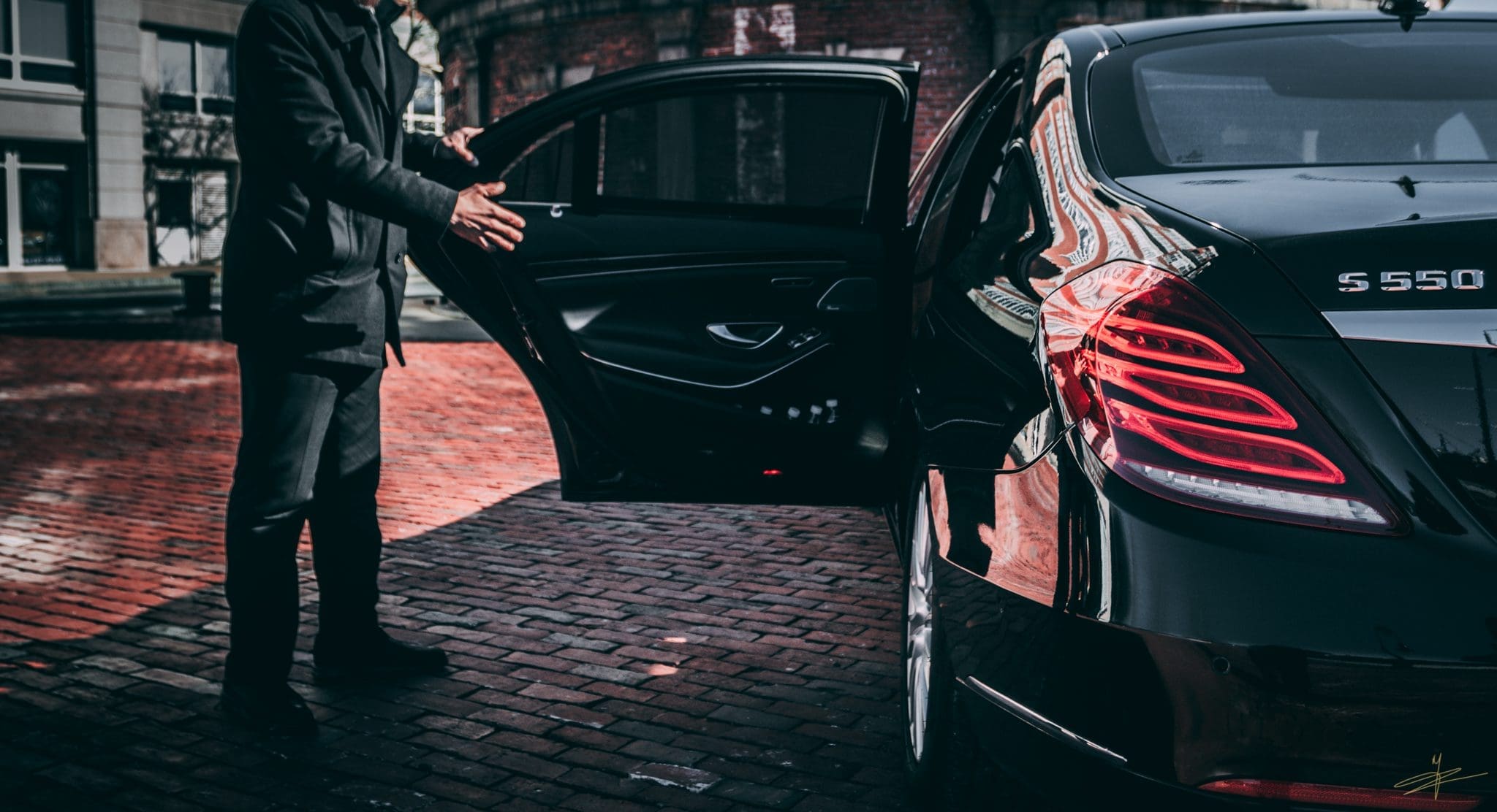 IMG_0167 A uniformed chauffeur assisting a passenger by opening the rear door of a luxury black sedan on a brick driveway for a Special Event Limousine service